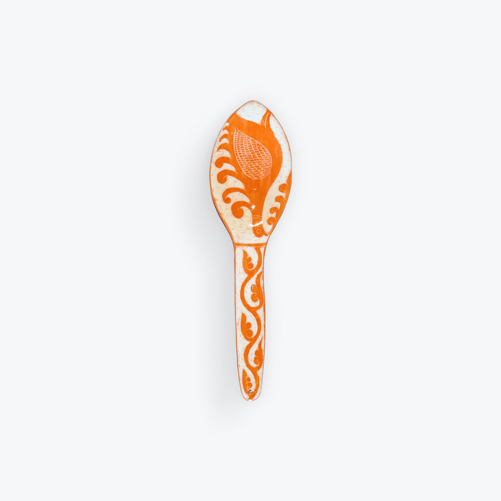 An orange and white decorative spoon with intricate hand-painted floral and scroll patterns, shown against a plain light gray background.