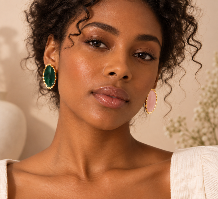 A close-up of a woman wearing large oval drop earrings with green and pink  in gold-tone settings, against a warm beige background.