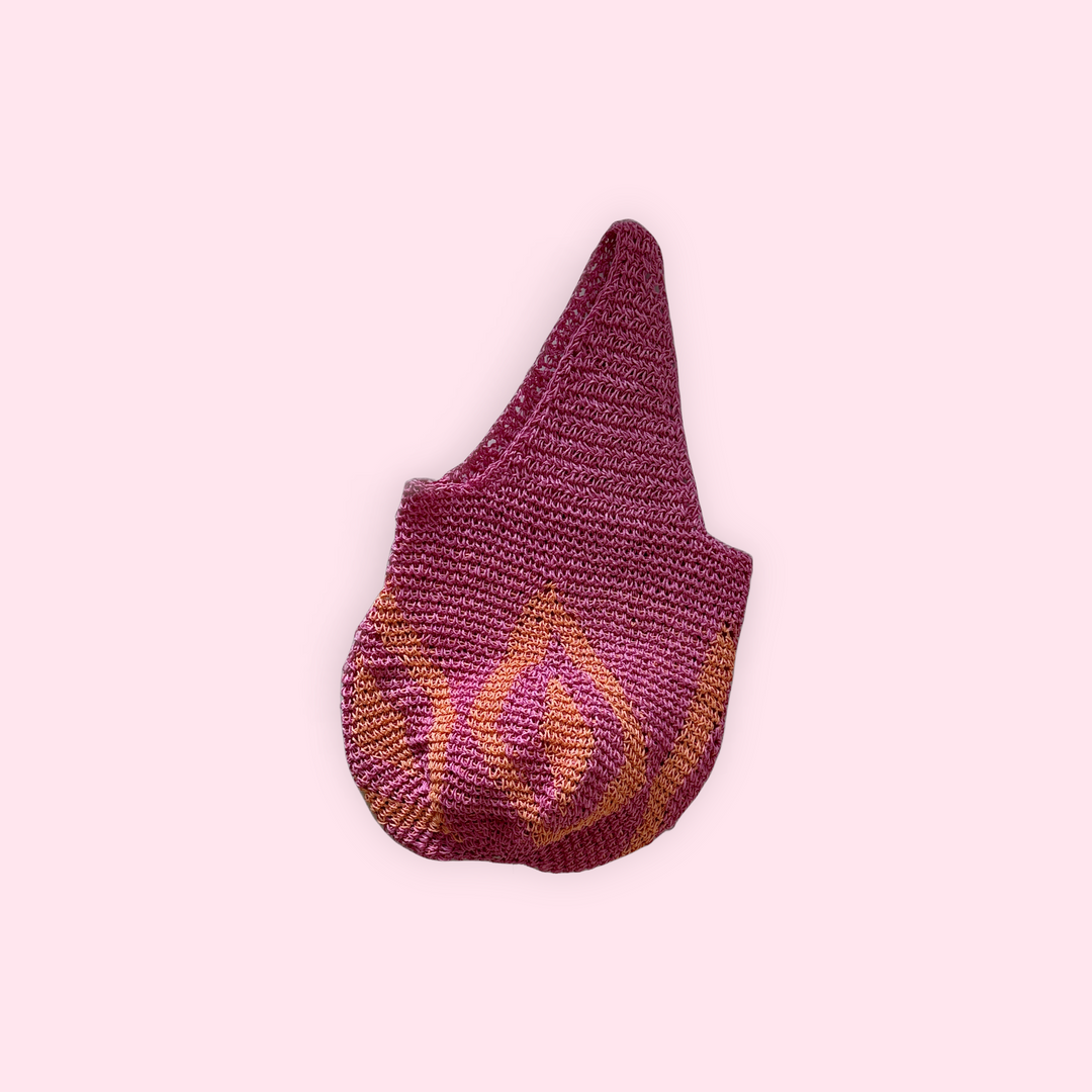 A single hand-knit slip-on sock in a deep magenta-purple color with a geometric diamond pattern in orange and yellow, shown against a solid pale pink background.