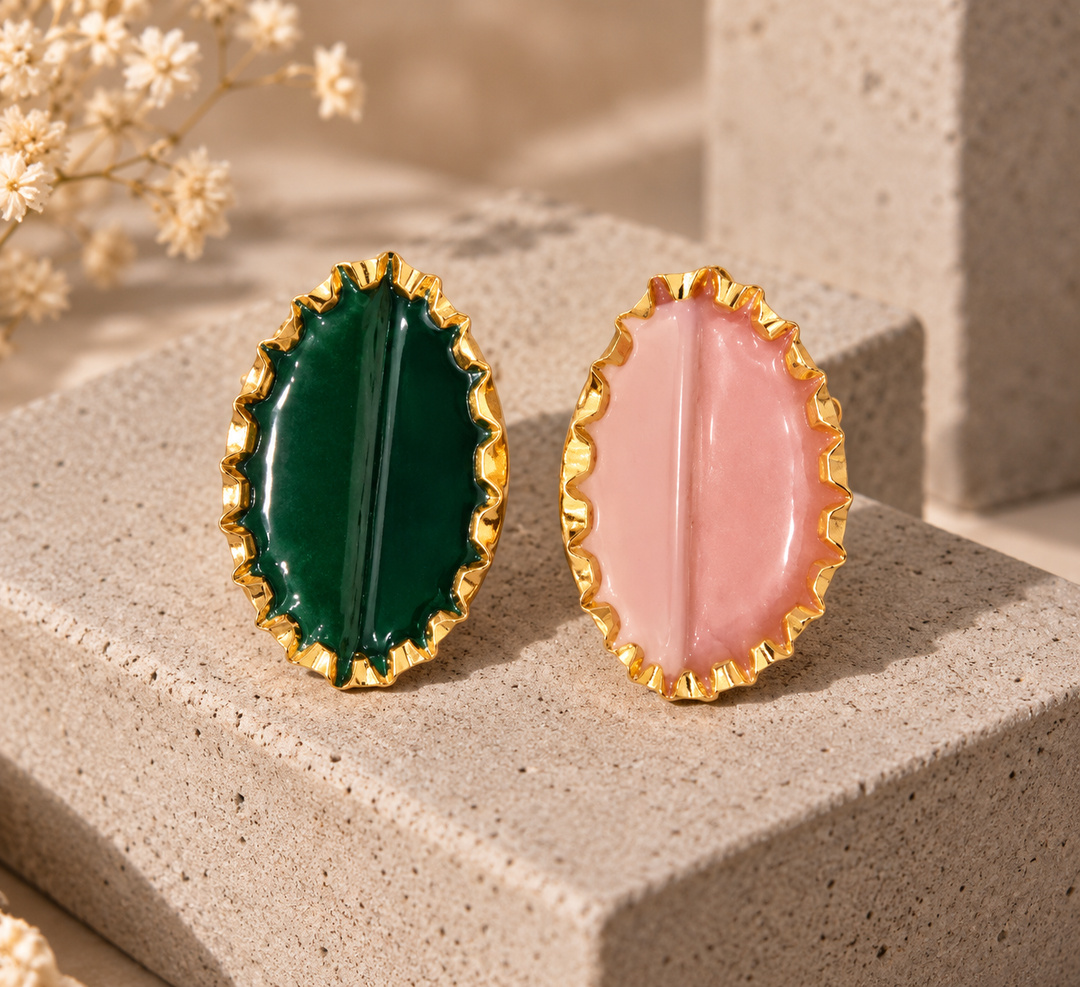 Two oval gold-tone earrings displayed side by side on a textured beige stone block. One earring features a deep emerald green center, and the other features a soft pink center.
