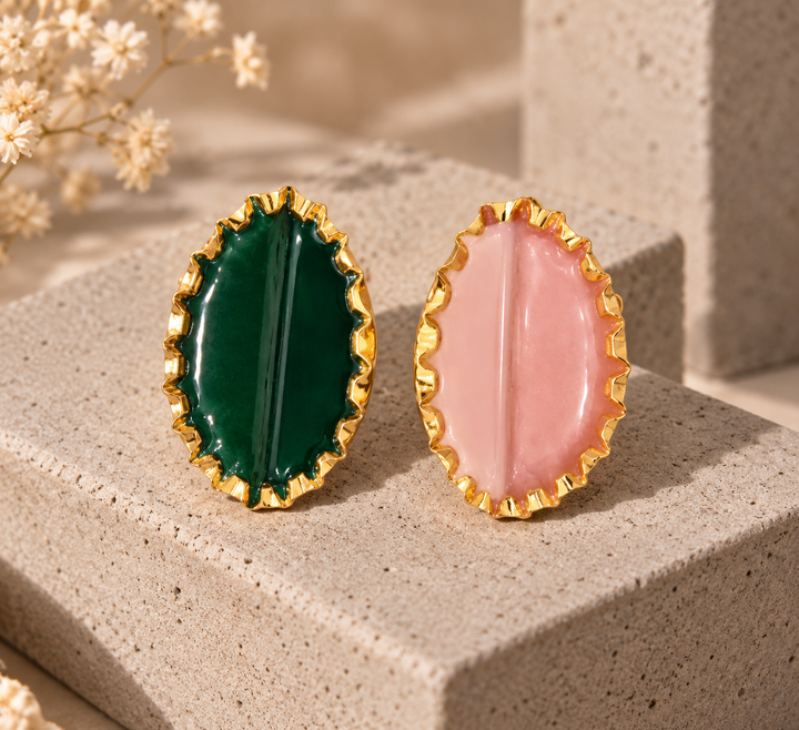 Two oval gold-tone earrings displayed side by side on a textured beige stone block. One earring features a deep emerald green center, and the other features a soft pink center.