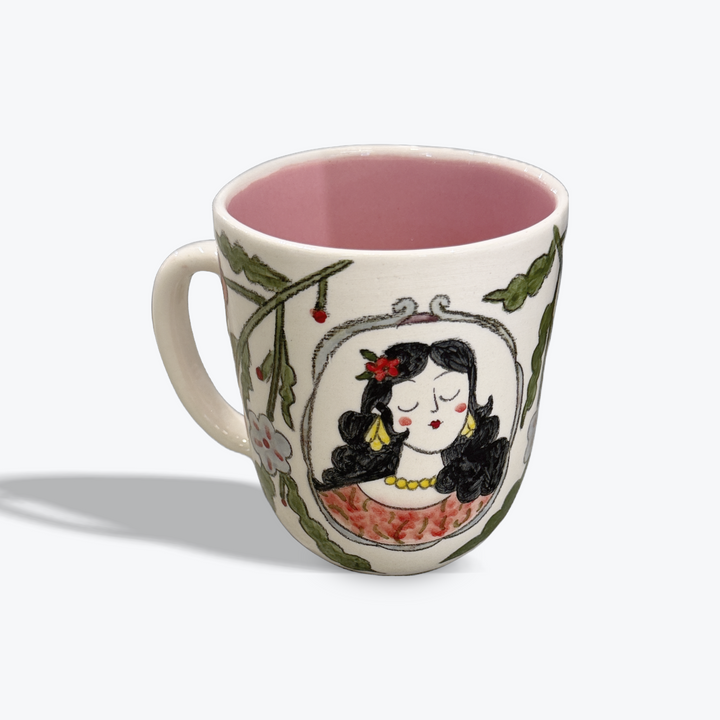 A ceramic mug with a pink interior and exterior illustration of a woman in a mirror surrounded by green leaves, on a light gray background.