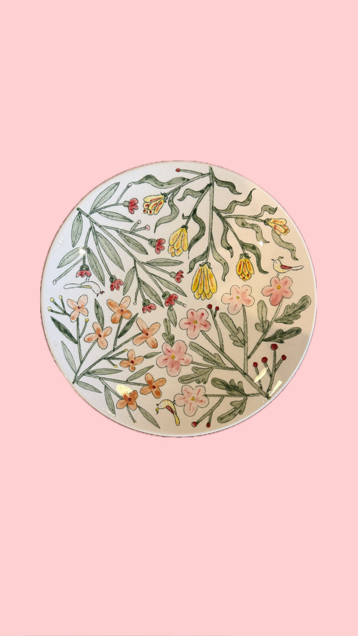 A round ceramic plate with a hand-painted botanical pattern of green leaves, yellow flowers, and pink blossoms on a cream background, set against a solid pink backdrop.