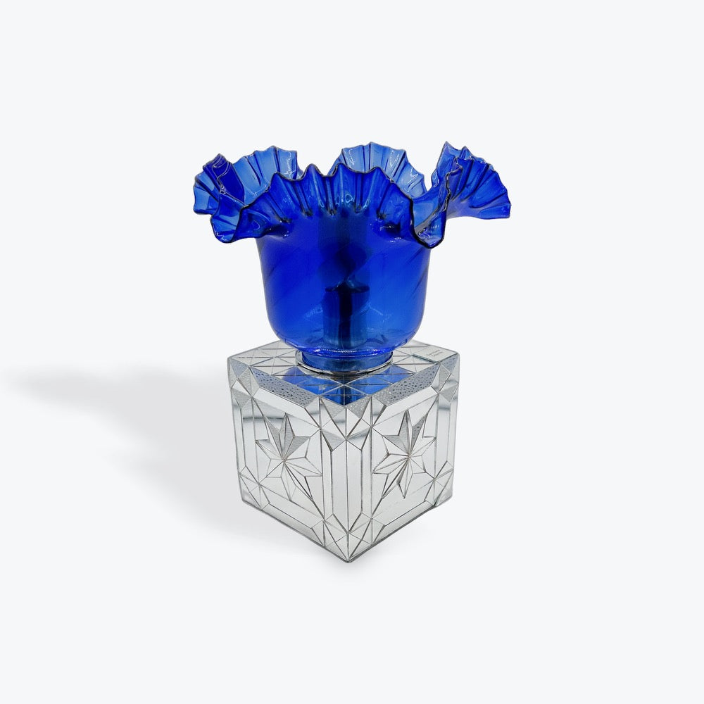 A cobalt blue ruffled glass vase or cup displayed on a clear faceted glass pedestal against a light gray studio background.