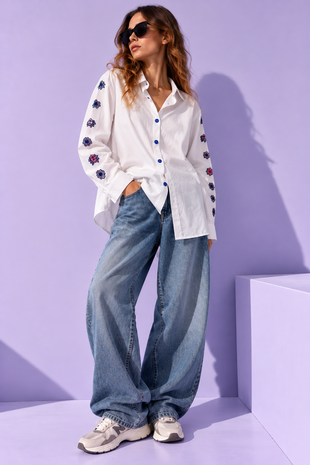 Model wearing a ZAAK white oversized shirt with hand-embroidered floral sleeves and blue buttons, styled with wide-leg denim jeans against a lavender background.