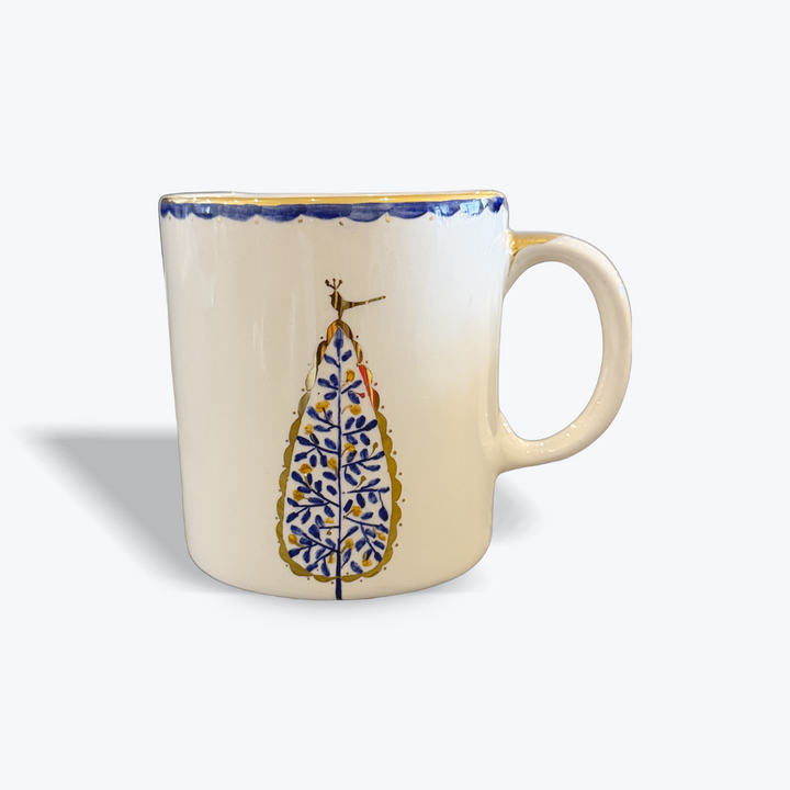 An off-white ceramic mug with a blue and gold hand-painted tree design on the front and a blue patterned rim, shown on a light gray background.
