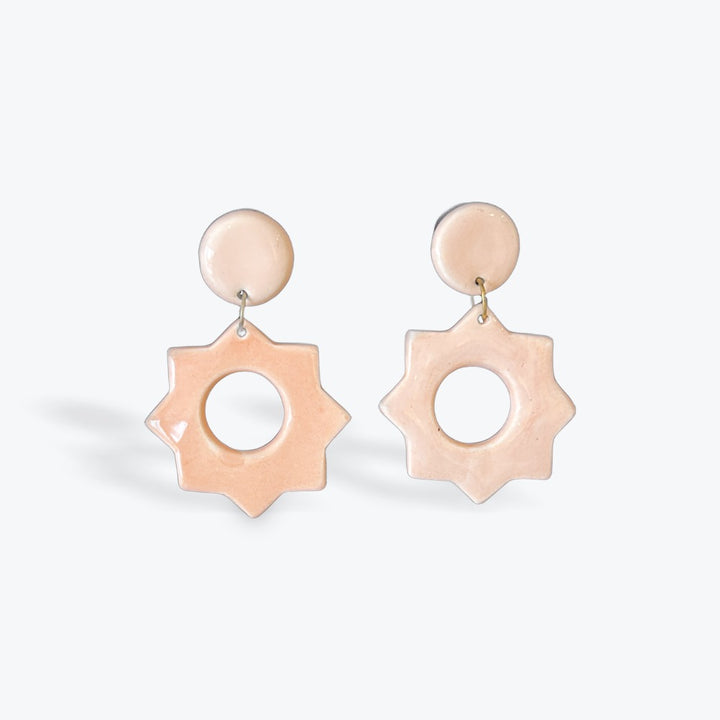 A pair of statement drop earrings featuring a circular pale pink top and a starburst-shaped dangling piece with a cutout center, shown on a light gray background.