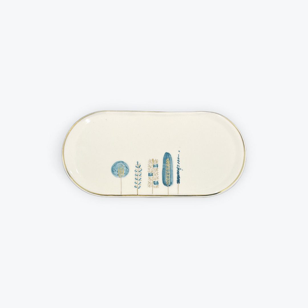 An oval ceramic serving tray with a cream-colored glaze, featuring a delicate gold rim and a minimalist blue and white botanical illustration along the bottom center.