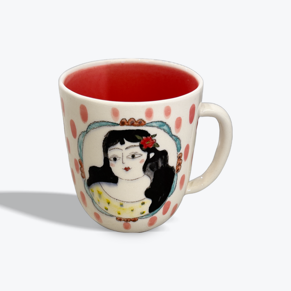 A ceramic coffee mug with a cream-colored exterior, decorated with pink polka dots and an illustration of a woman with black hair and a red flower in her hair. The mug features a vibrant red interior and a white handle, shown on a light gray background.