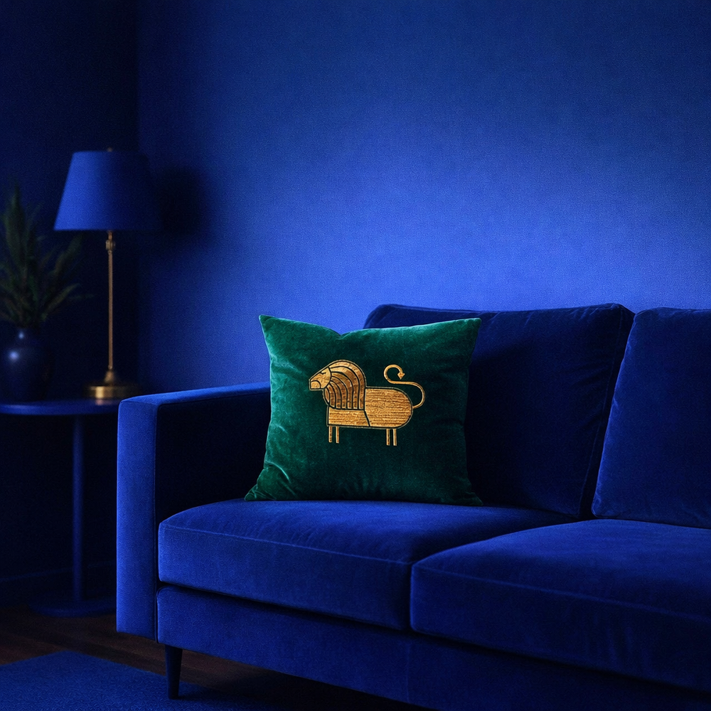 A dark green velvet throw pillow with a gold embroidered lion design, styled on a deep blue velvet sofa in a dimly lit room with a blue wall and a side table with a lamp.
