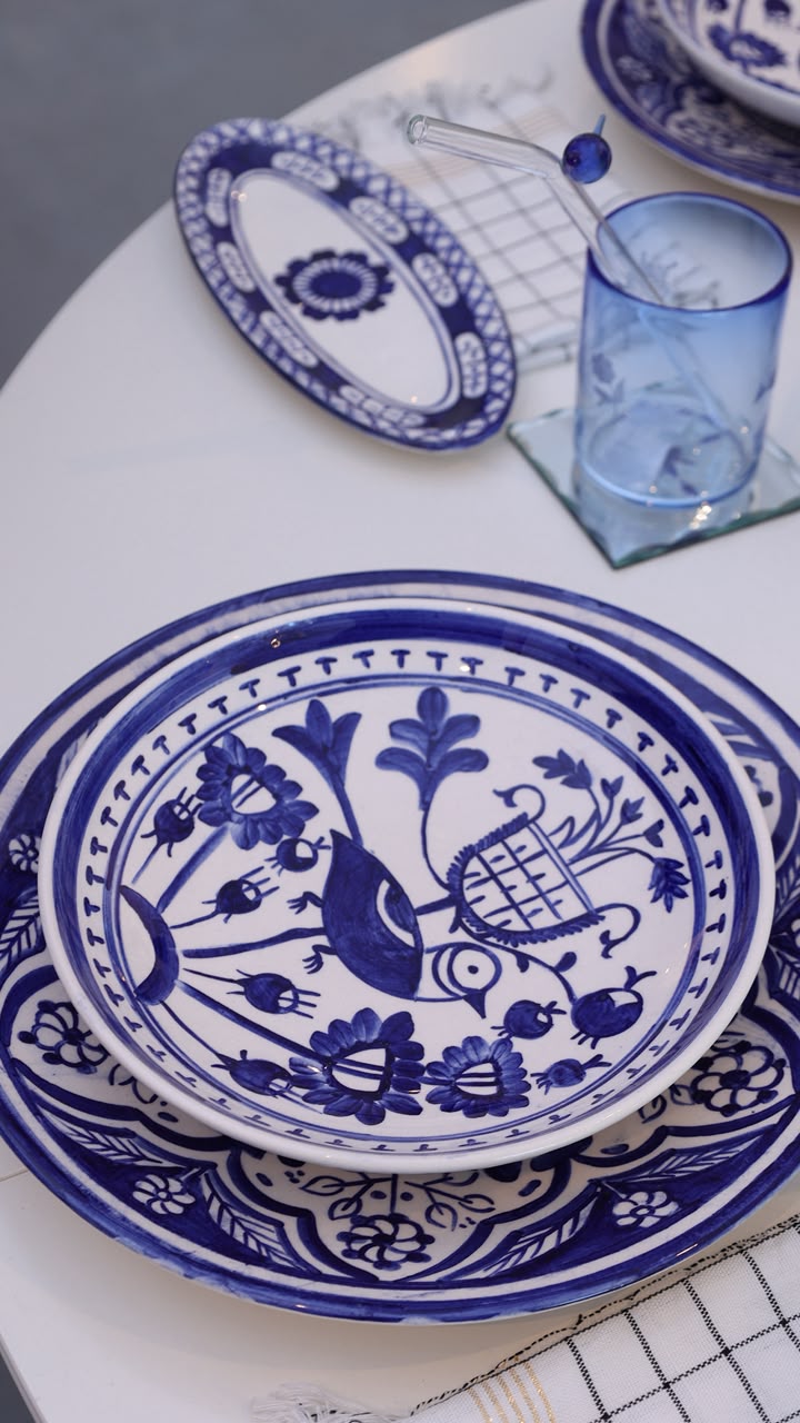 Blue and white ceramic tableware set on a white table, featuring a large floral-patterned plate, a matching charger plate, a smaller oval plate, and a translucent blue glass tumbler.