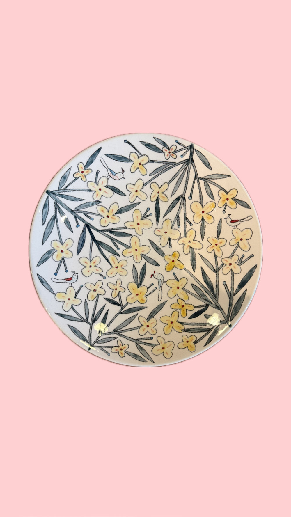 A round ceramic plate featuring a hand-painted floral pattern of yellow flowers and green leaves on a white background, set against a solid pink backdrop.