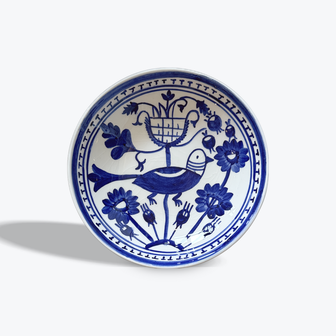 A round white ceramic bowl with a blue hand-painted design featuring a bird, flowers, and a basket, photographed on a light gray background.