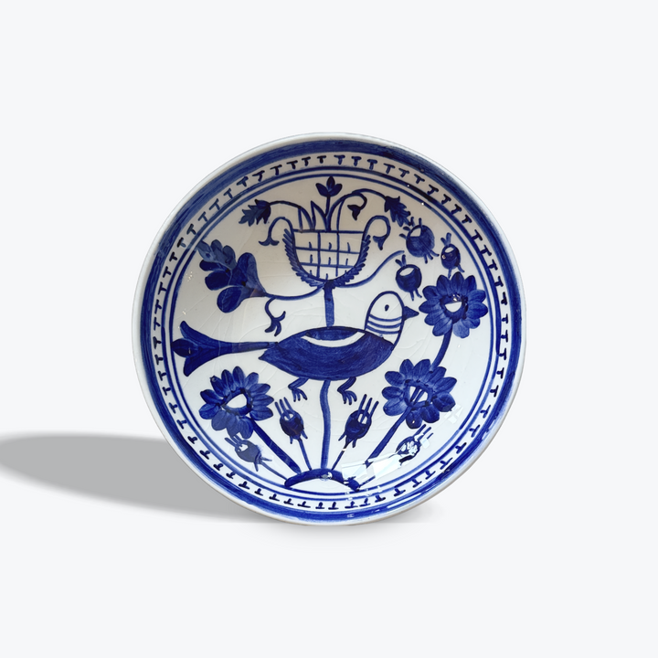 A round white ceramic bowl with a blue hand-painted design featuring a bird, flowers, and a basket, photographed on a light gray background.