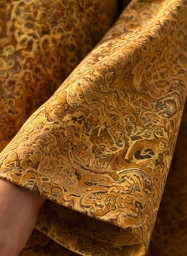A close-up of a textured fabric sleeve or garment section featuring an intricate golden-yellow and brown abstract pattern, with a hand holding the edge to show drape and texture.