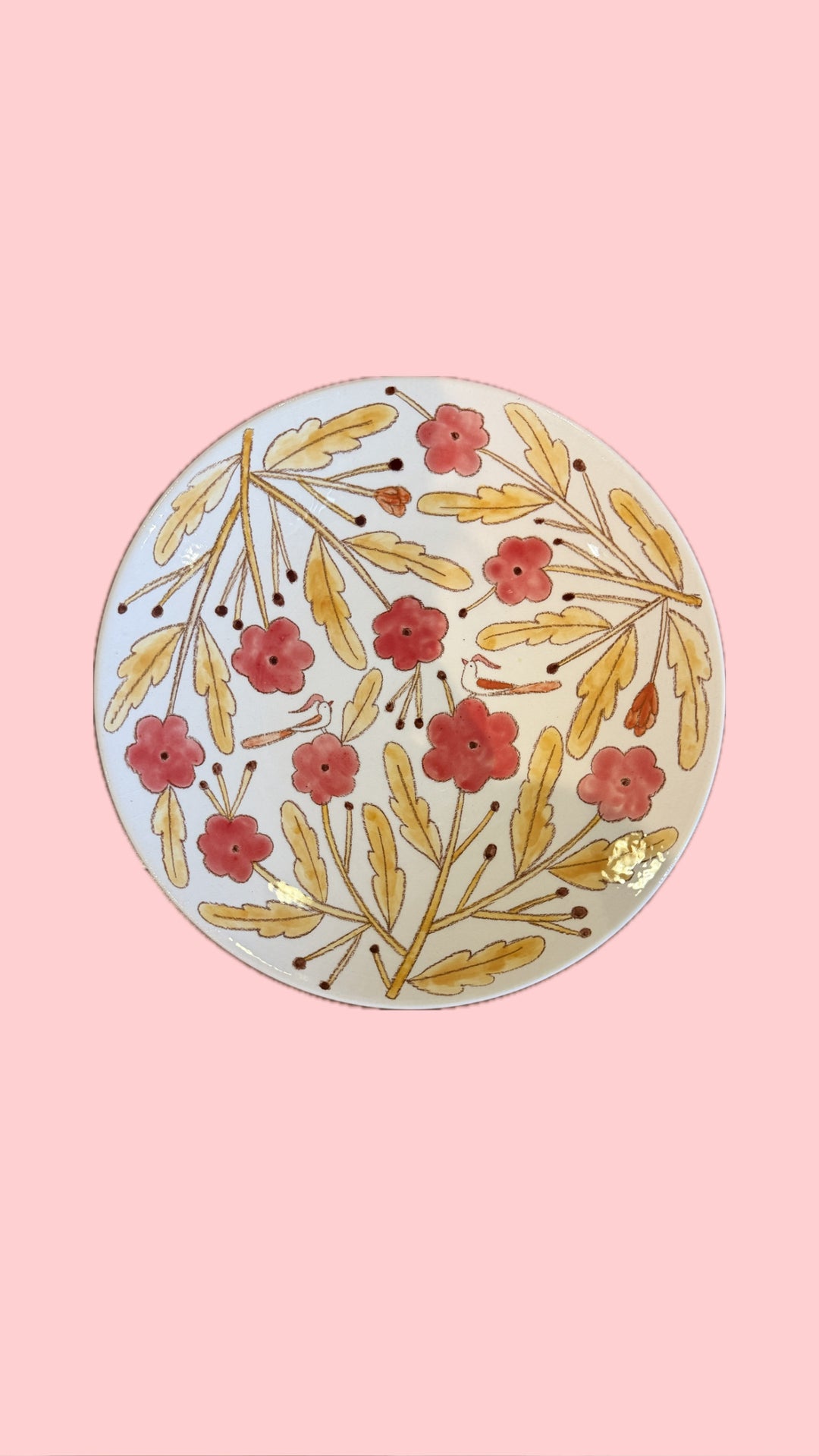 A round ceramic plate with a hand-painted floral pattern of red flowers and gold leaves on a white background, set against a solid pink backdrop.