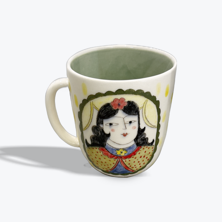 A ceramic mug with a handle, featuring a hand-painted illustration of a stylized woman's face on the front, set against a plain white background.