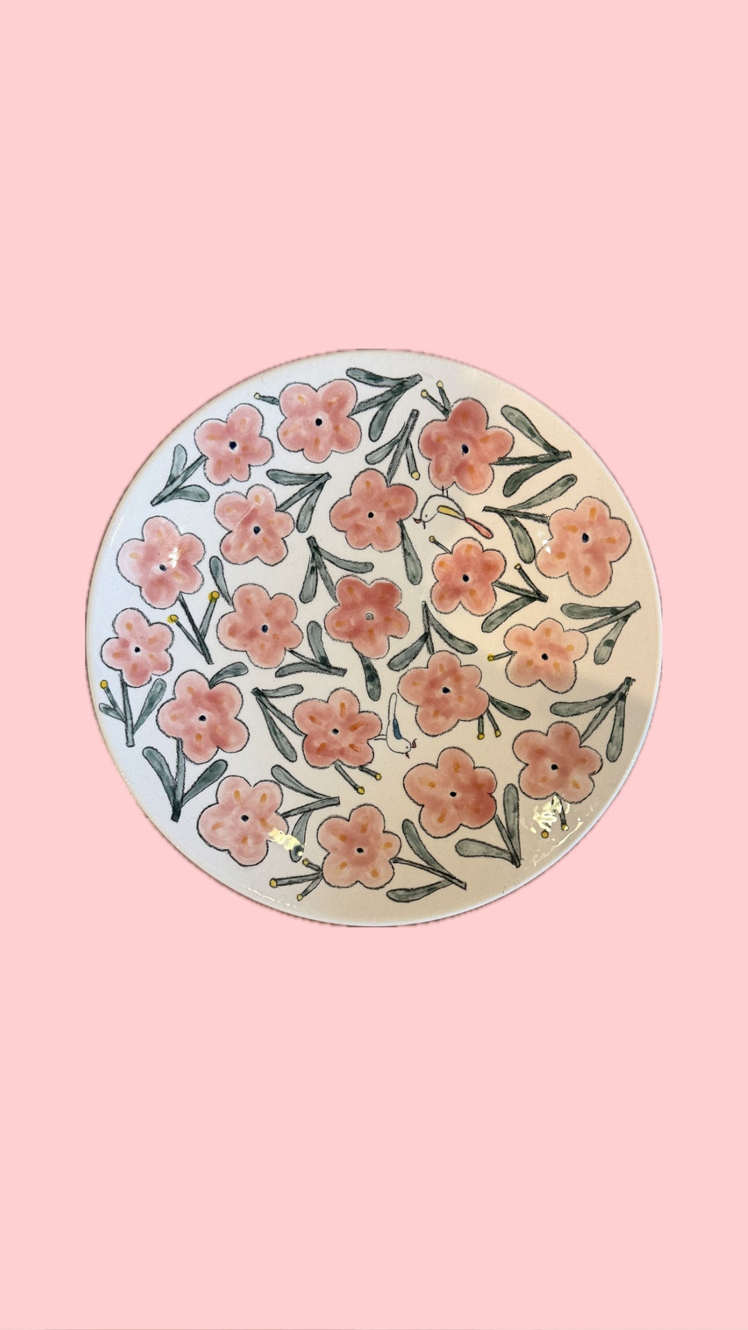 A round ceramic plate with a hand-painted floral pattern of pink and orange flowers with green leaves on a cream background, set against a solid pink backdrop.