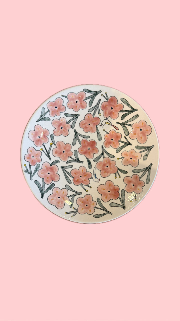 A round ceramic plate with a hand-painted floral pattern of pink and orange flowers with green leaves on a cream background, set against a solid pink backdrop.