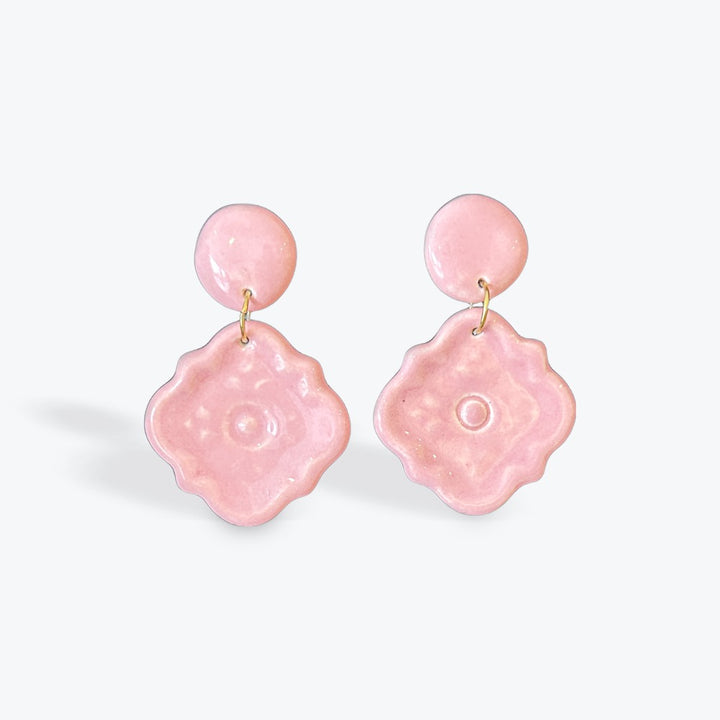 A pair of light pink dangling earrings with round studs and textured, flower-like drop charms, shown on a clean white background.