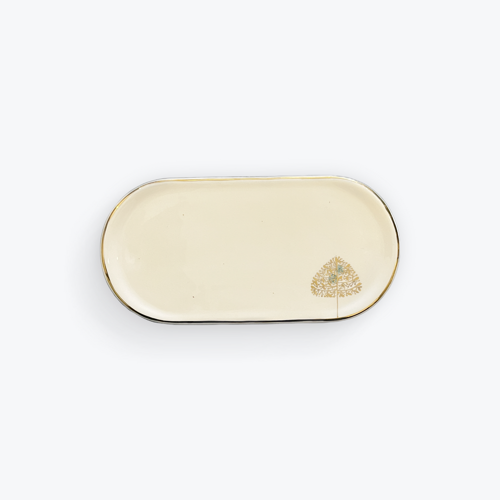 An oval ceramic serving tray with a cream-colored glaze, a thin gold rim, and a small gold tree illustration in the bottom right corner, set against a plain white background.