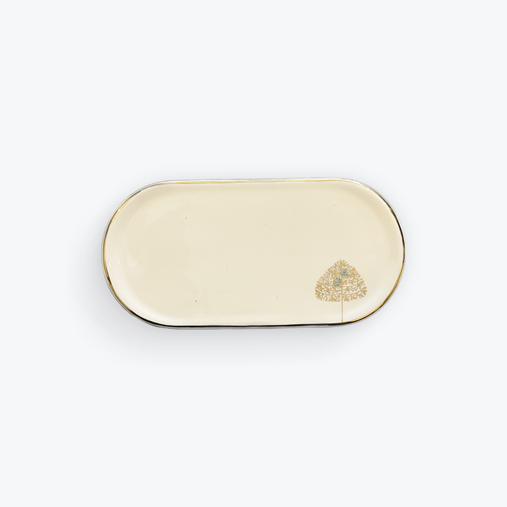 An oval ceramic serving tray with a cream-colored glaze, a thin gold rim, and a small gold tree illustration in the bottom right corner, set against a plain white background.
