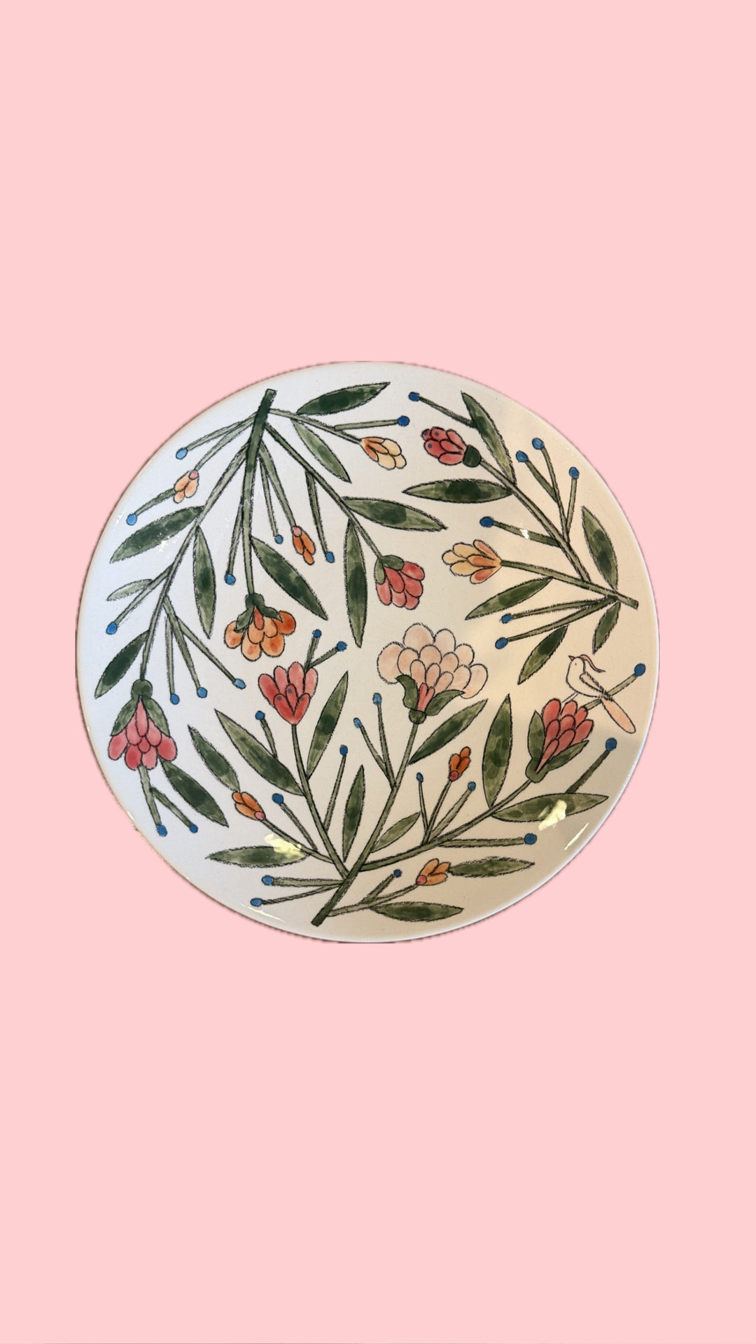 A round ceramic plate with a hand-painted botanical pattern of green leaves and pink, orange, and white flowers on a cream background, set against a solid pink backdrop.