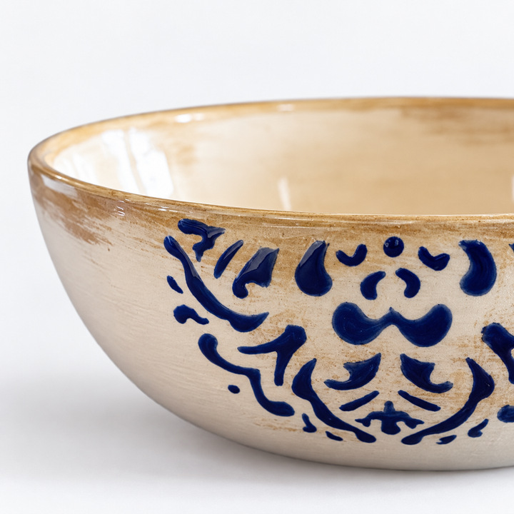 A close-up of a ceramic bowl with a warm beige glaze, featuring a rustic brown rim and a dark blue hand-painted floral pattern on the side.