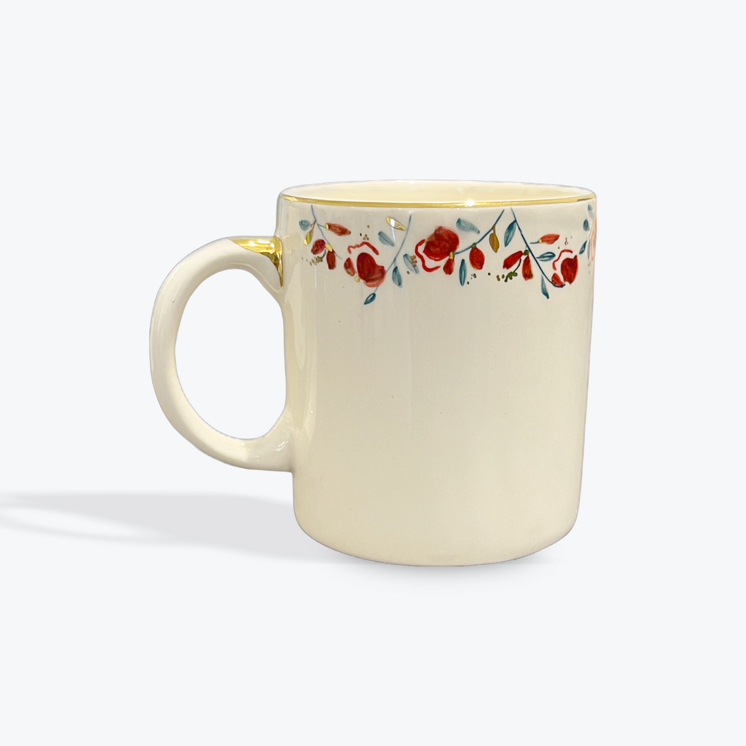 An off-white ceramic mug with a delicate floral border pattern in red, blue, and green, featuring a gold accent on the rim and handle, set against a light gray background.