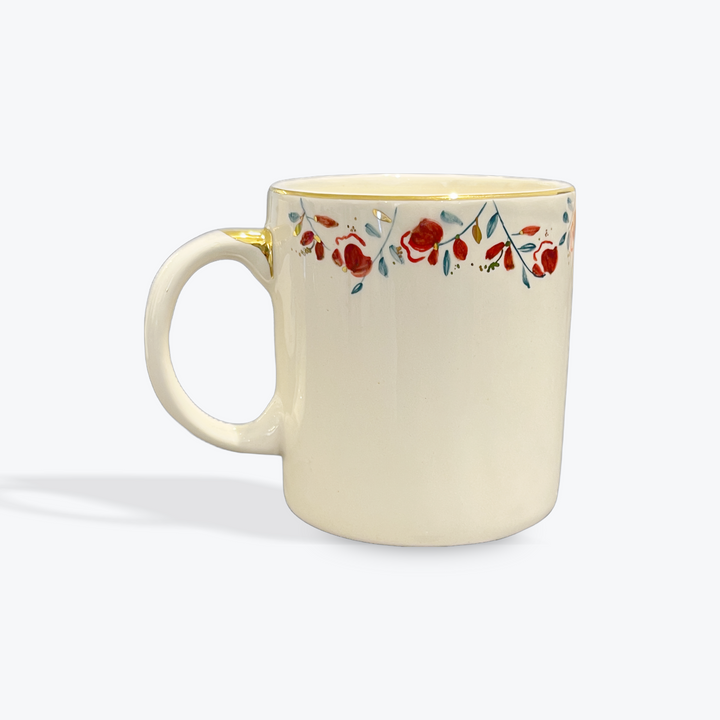 An off-white ceramic mug with a delicate floral border pattern in red, blue, and green, featuring a gold accent on the rim and handle, set against a light gray background.