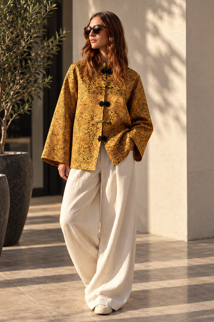 A woman wearing a mustard yellow patterned jacket with black buttons and wide-leg cream trousers, standing outdoors in front of a beige wall with shadows.