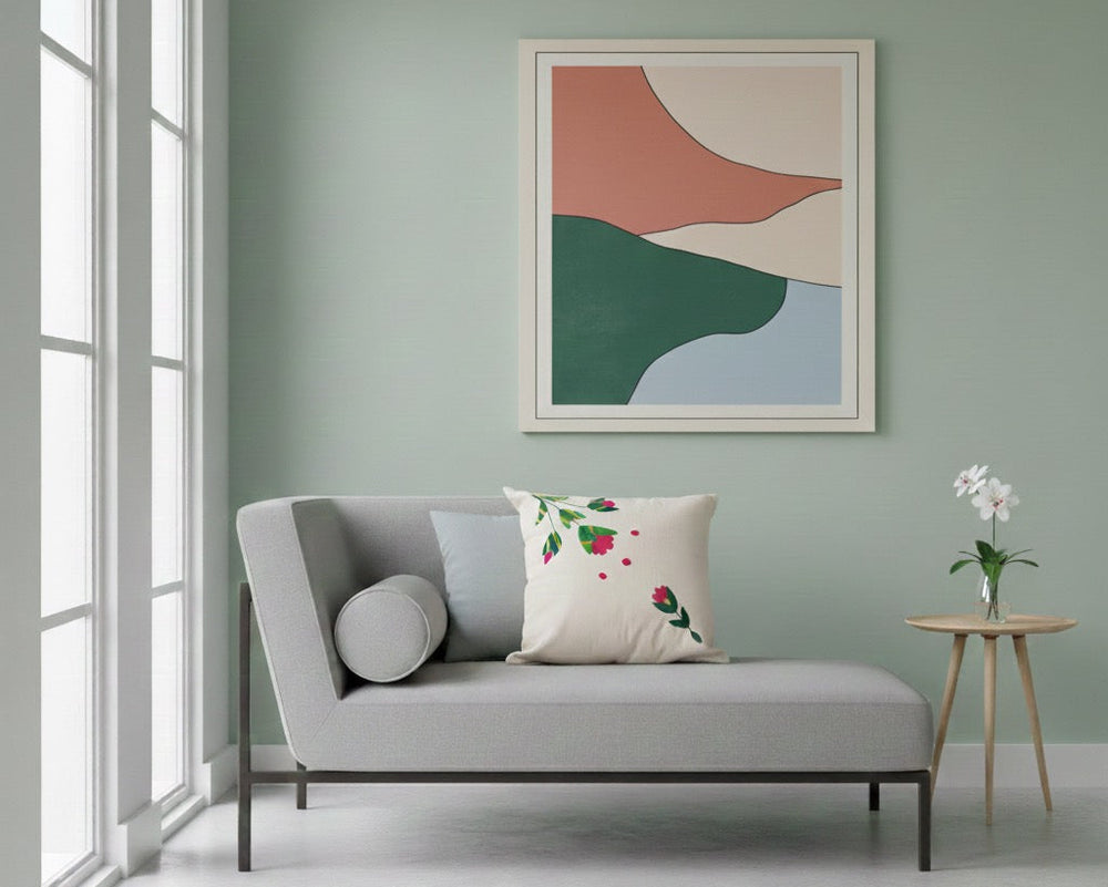 A modern gray chaise lounge with a floral accent pillow, set in a minimalist living room with a sage green wall and an abstract framed art print above.