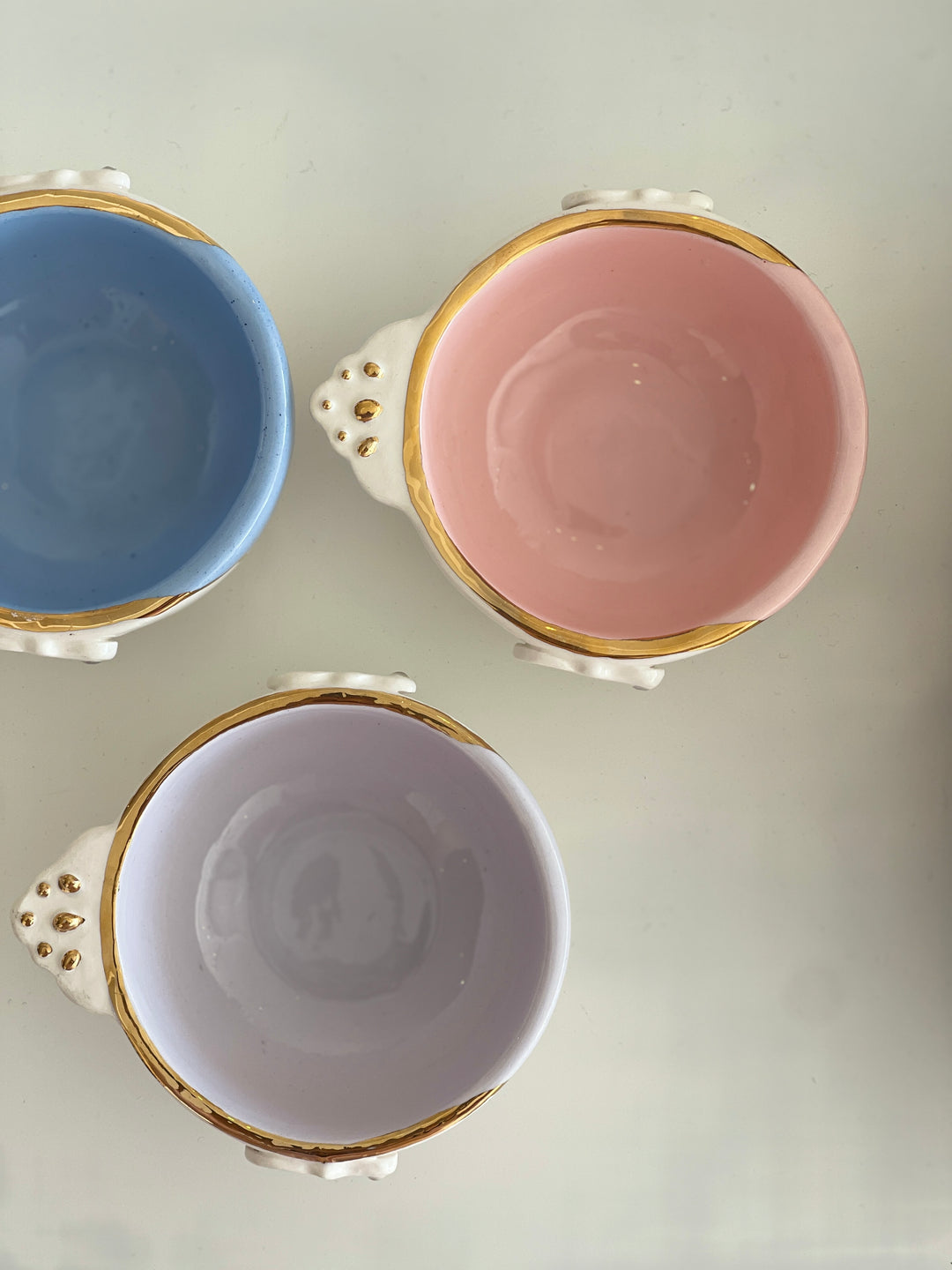 Three ceramic cups with gold trim in blue, pink, and white on a light gray background