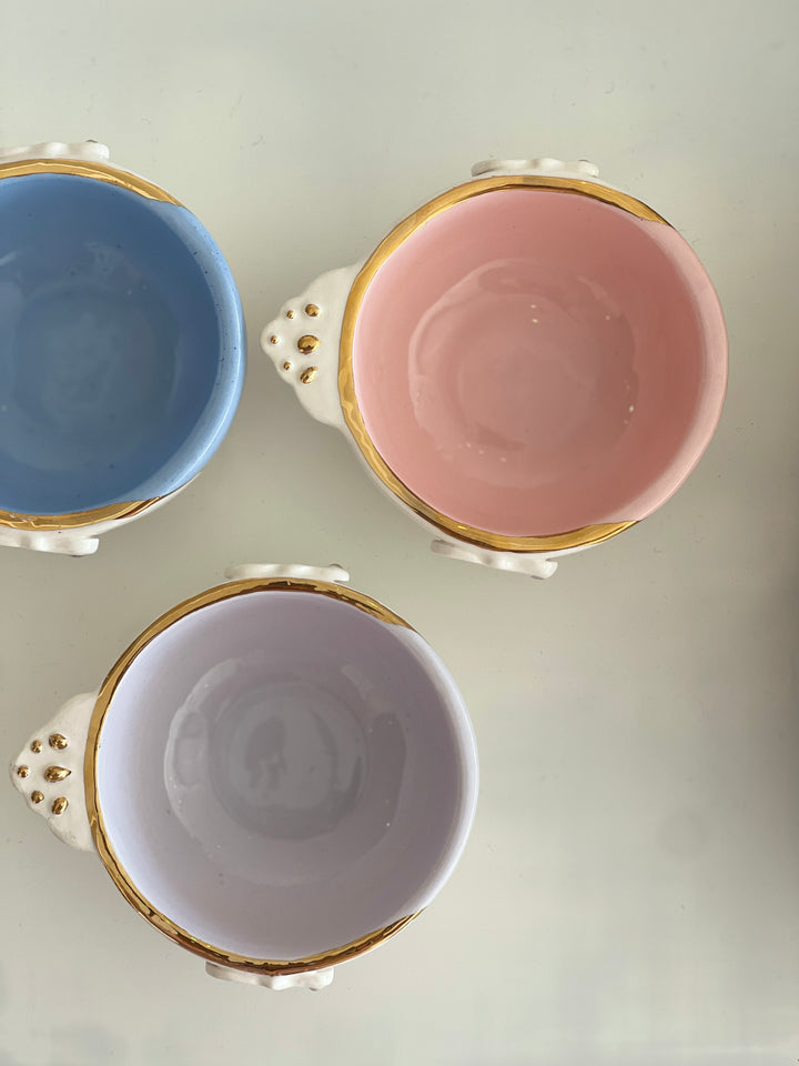 Three ceramic cups with gold trim in blue, pink, and white on a light gray background