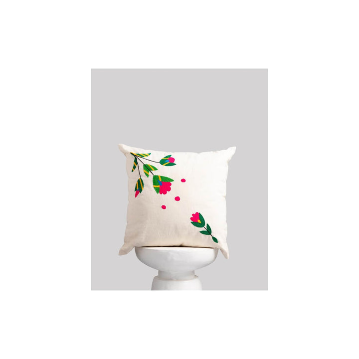 An off-white square throw pillow with embroidered pink flowers and green leaves, displayed on a white pedestal against a light gray background.