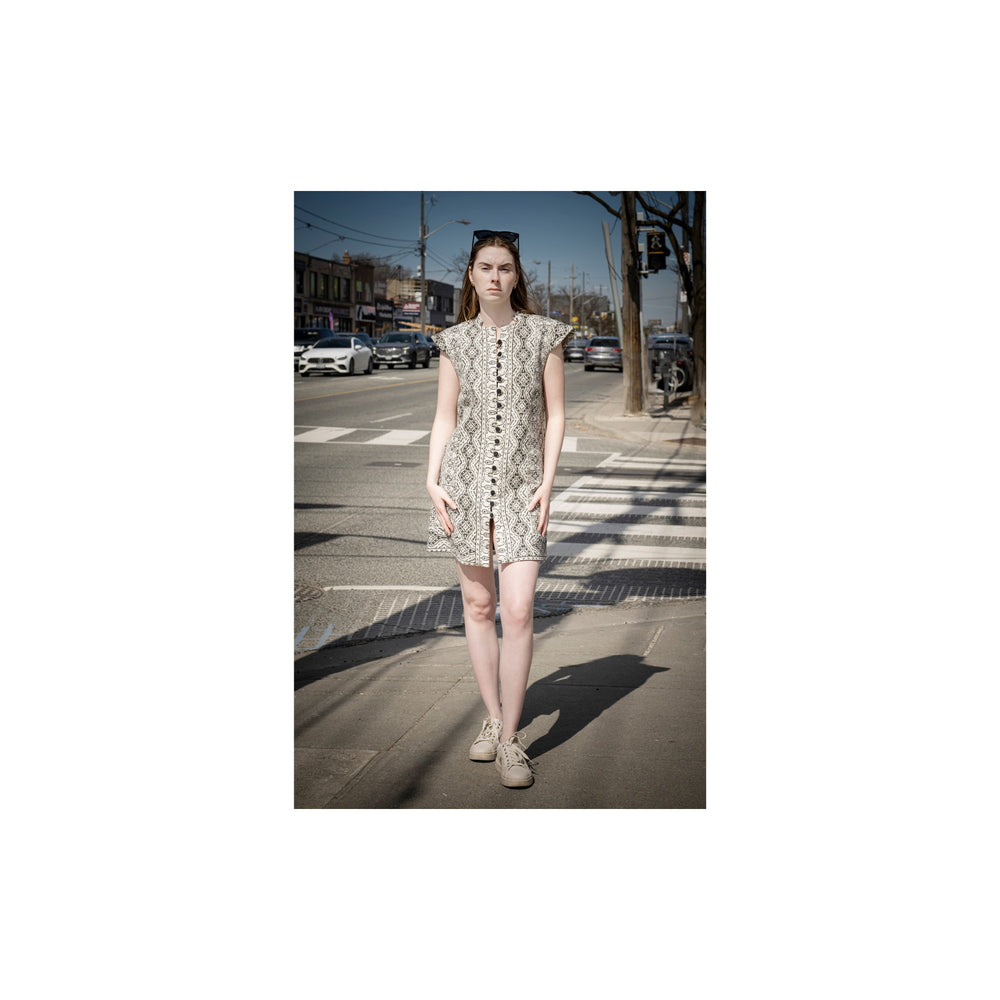 A woman stands on a city street crosswalk wearing a sleeveless beige and black snakeskin-pattern mini dress with a front slit and matching belt, paired with beige sneakers.