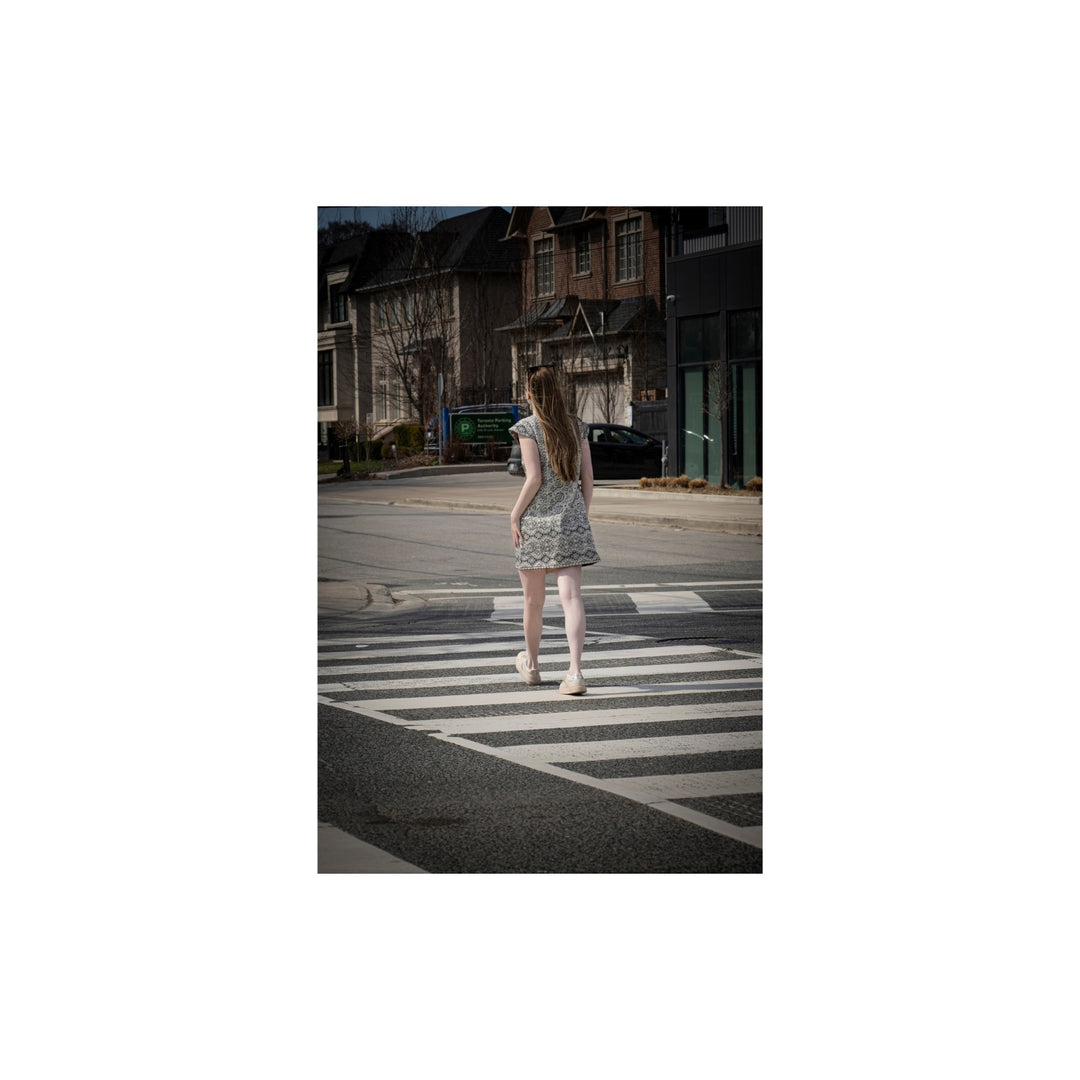 A woman in a light patterned dress walks across a city crosswalk, viewed from behind, with buildings and a street in the background.