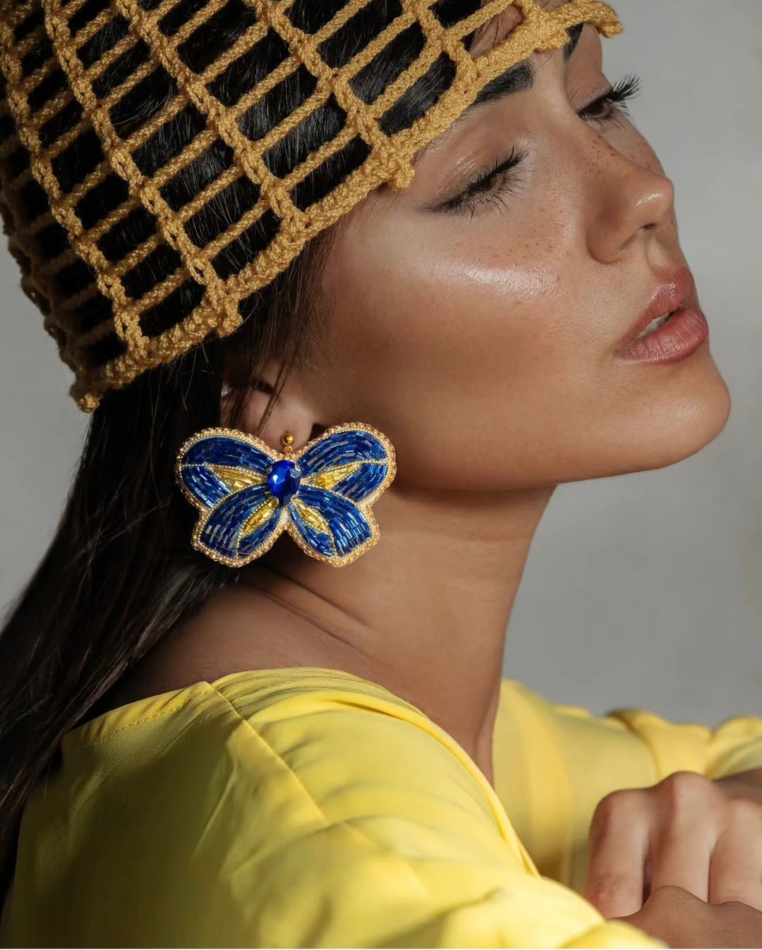 A close-up side profile of a woman wearing a yellow top, a yellow woven hat, and a large butterfly-shaped earring in blue and yellow with gold detailing.