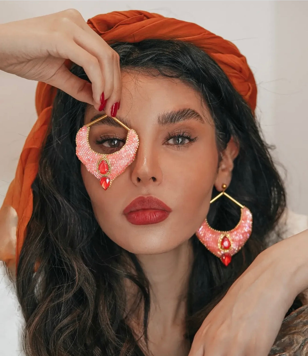 A close-up lifestyle shot of a model wearing large, bohemian-style gold-tone earrings with pink beaded crescent shapes and red teardrop gems, along with a matching rust-orange headwrap.