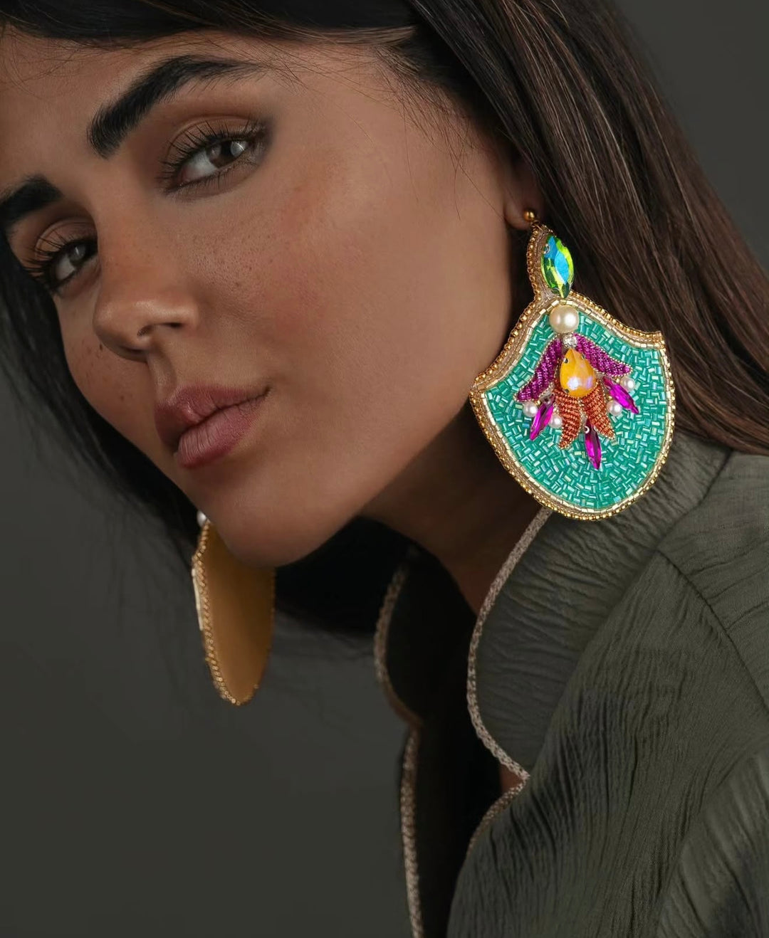 A close-up of a woman wearing a large, ornate turquoise and gold statement earring with colorful beadwork and a matching gold disc earring.