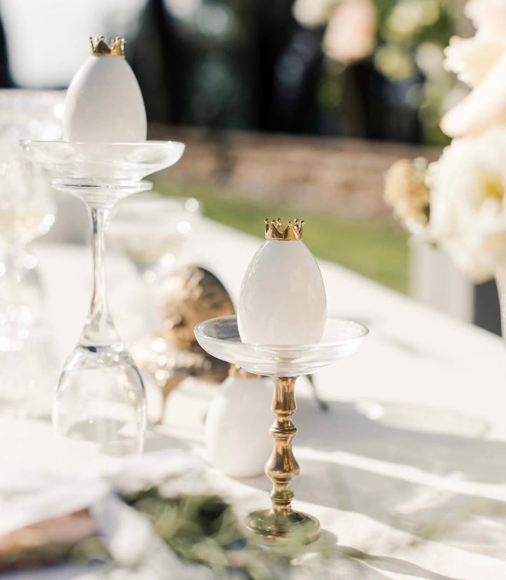 White decorative eggs with small gold crowns displayed on clear and gold pedestal stands on a softly lit table setting.