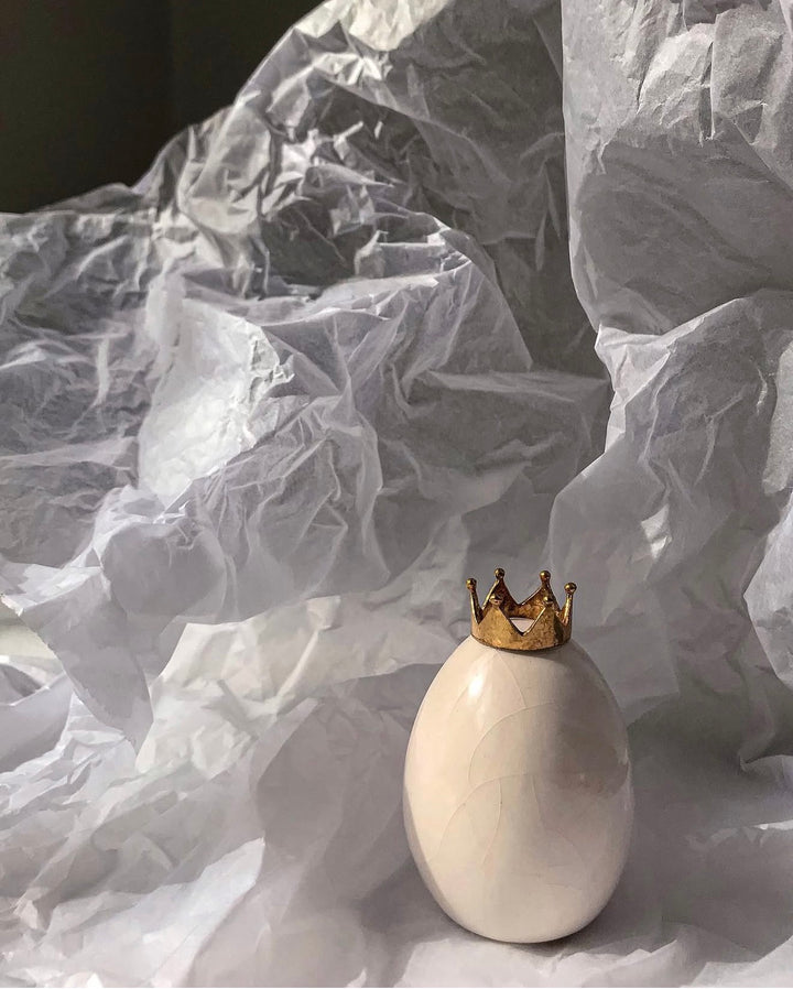 A glossy white ceramic egg-shaped vase with a small gold crown on top, set against a backdrop of crumpled white tissue paper.