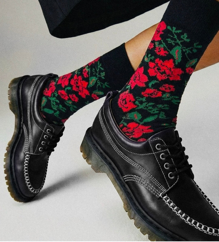 A close-up of a person's legs wearing black leather moc-toe lace-up shoes and black crew socks with a vibrant red rose and green leaf floral pattern, set against a neutral light gray background.