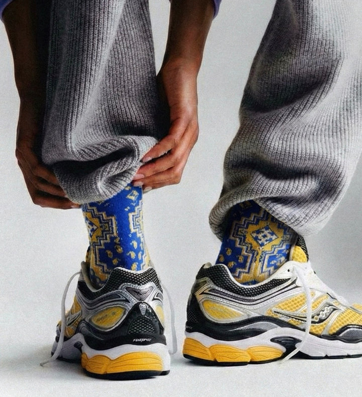 A close-up of a person's lower legs and feet, wearing gray ribbed sweatpants, patterned blue and yellow socks, and chunky yellow, black, and white sneakers against a light gray background.