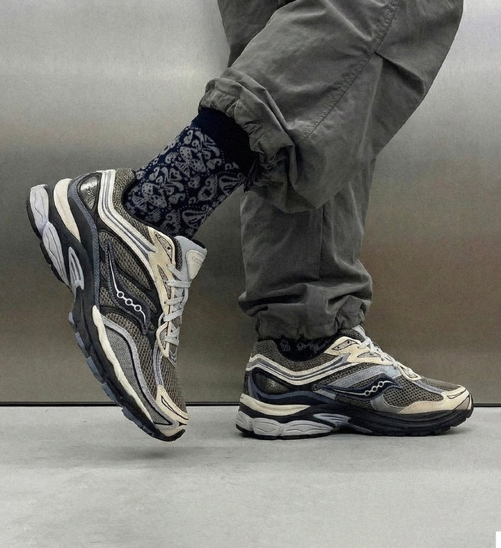 A person wearing gray cargo pants, patterned socks, and a pair of Saucony chunky sneakers in gray, black, and off-white, standing against a metallic gray background.