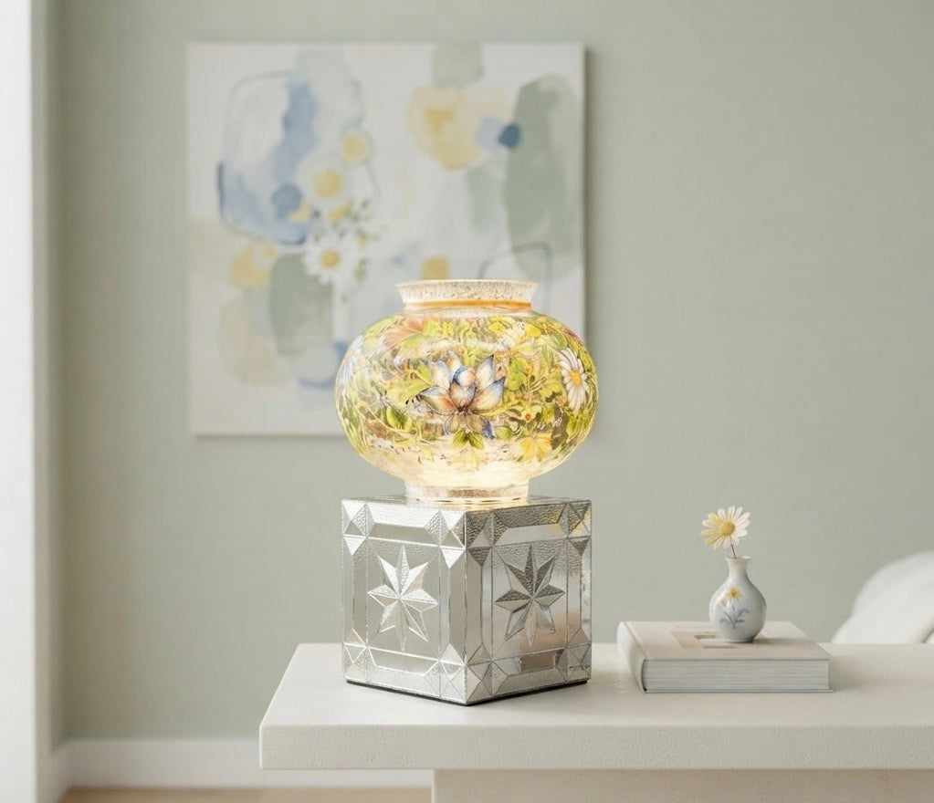 A decorative glass jar with a yellow floral pattern sits on a mirrored silver pedestal base, displayed on a white side table in a styled room setting.