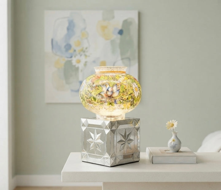 A decorative glass jar with a yellow floral pattern sits on a mirrored silver pedestal base, displayed on a white side table in a styled room setting.