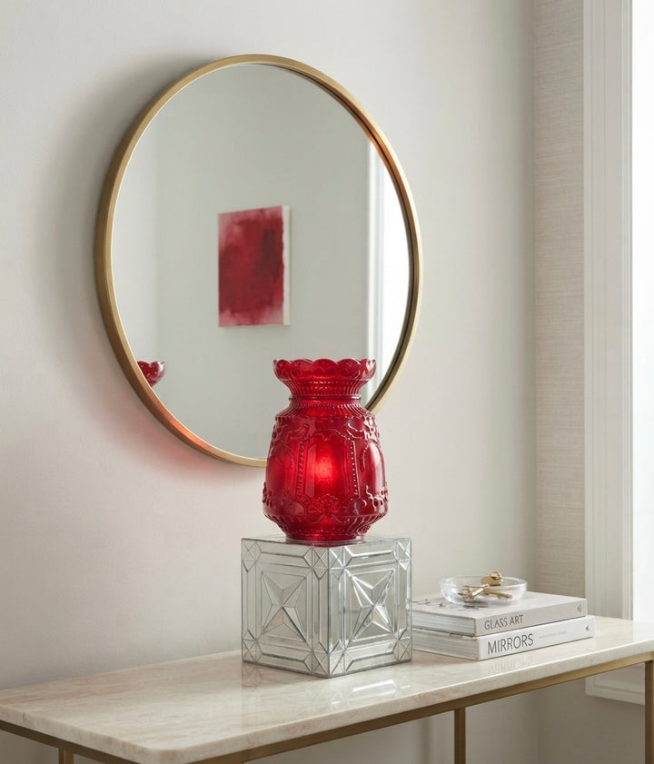 A round wall mirror with a gold frame hangs above a console table. On the table sits a red glass vase on a mirrored pedestal, next to a stack of books.