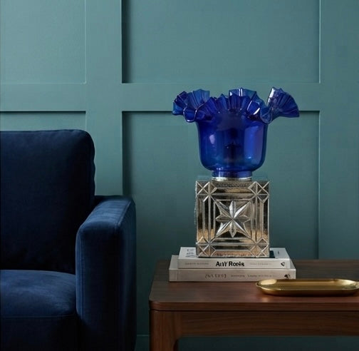 A cobalt blue ruffled glass vase on a mirrored geometric pedestal, styled on books atop a wooden side table next to a dark blue armchair against a teal paneled wall.