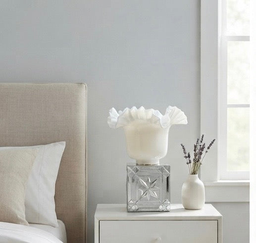 A white ruffled fabric-covered table lamp with a clear faceted glass base, displayed on a white nightstand next to a small white vase with dried lavender in a bedroom setting.